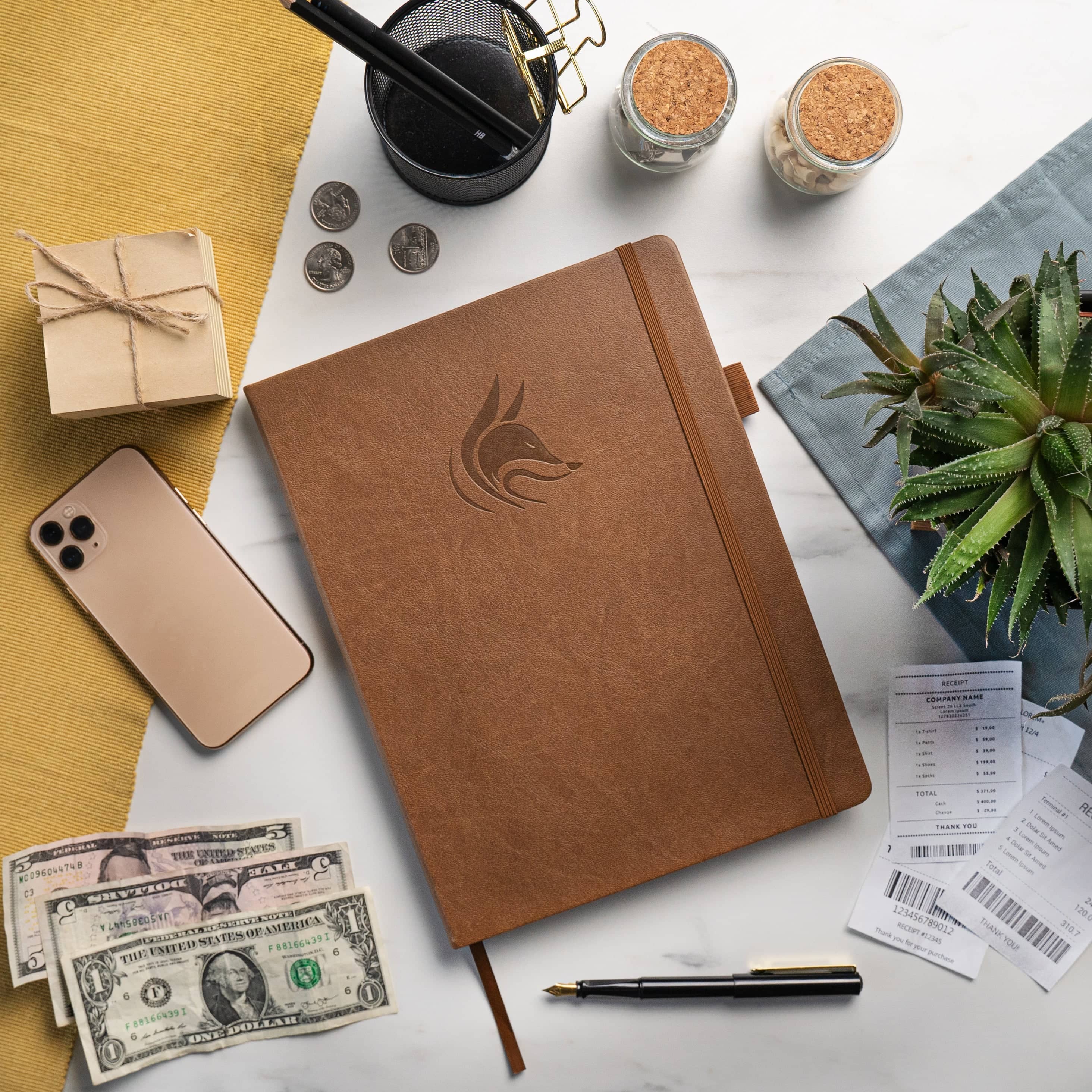 A brown textured leather planner or notebook with an embossed fox logo, styled in a flat-lay arrangement on a white marble surface alongside a smartphone, cash, receipts, a pen, and other desk accessories.