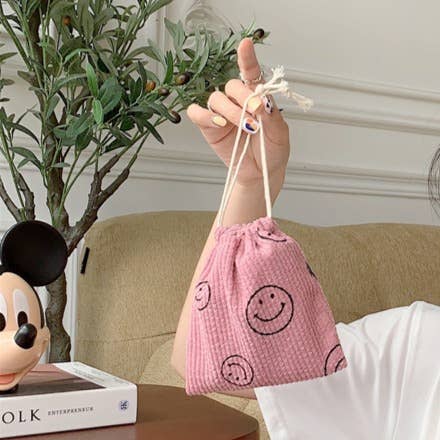 Pink drawstring bag with smiley face patterns held by a hand, with a plant and book in the background.