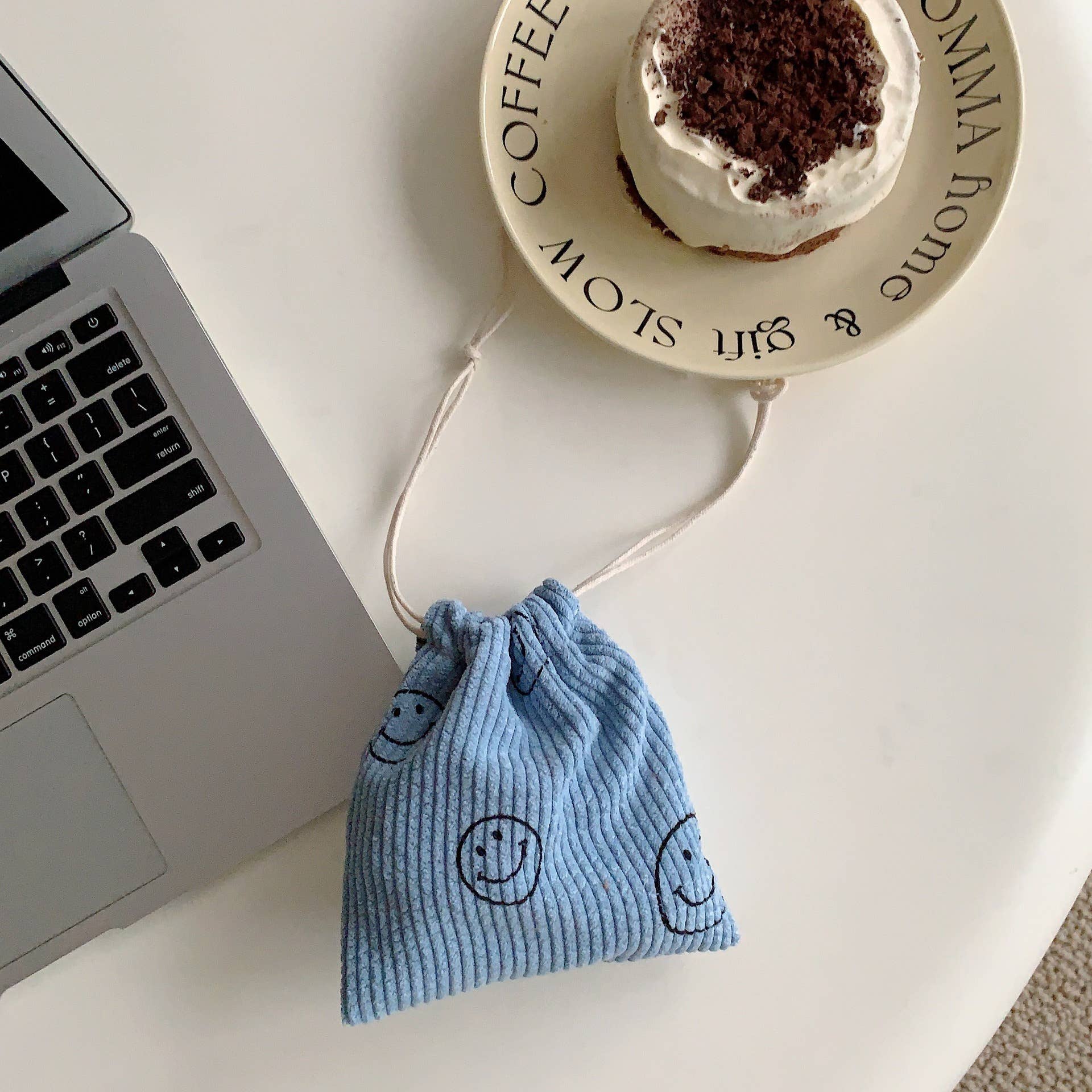 Blue drawstring bag with smiley faces next to a laptop and a cup of coffee on a white surface.