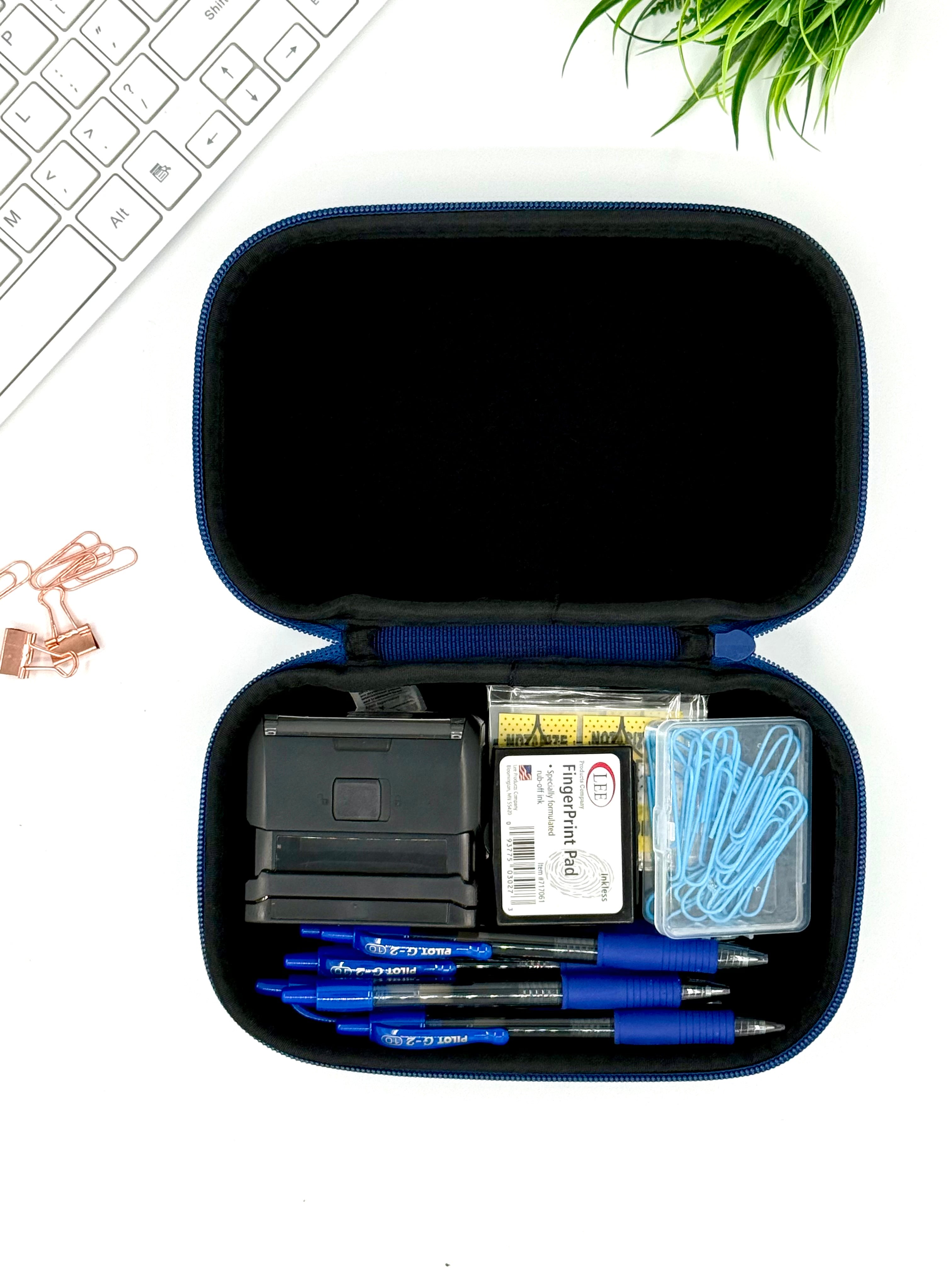 Open blue case containing stationery items on a white surface with a keyboard and plant in the background.