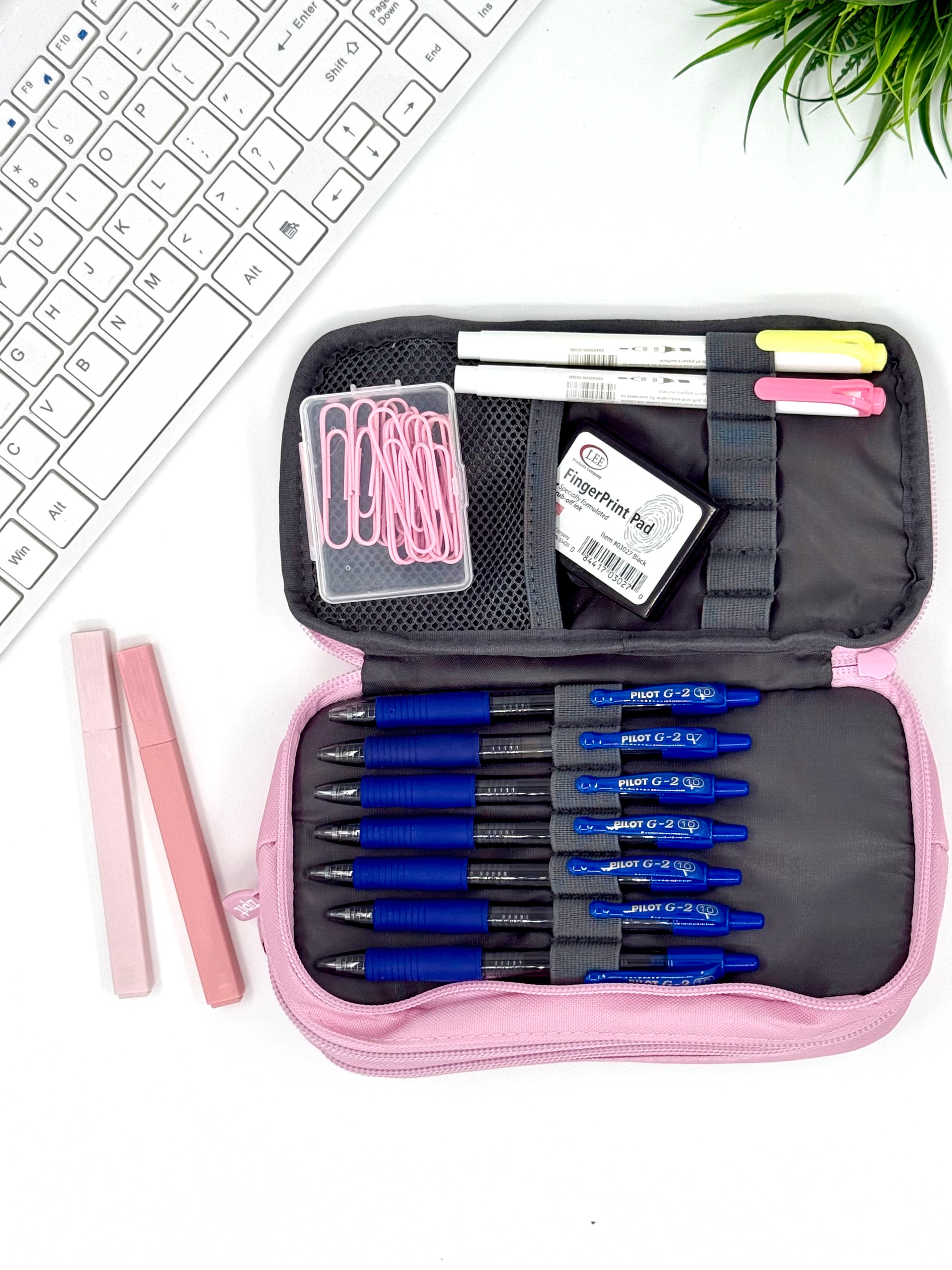 Pink pencil case with blue and black pens, a pink pen, and a white keyboard on a white background.