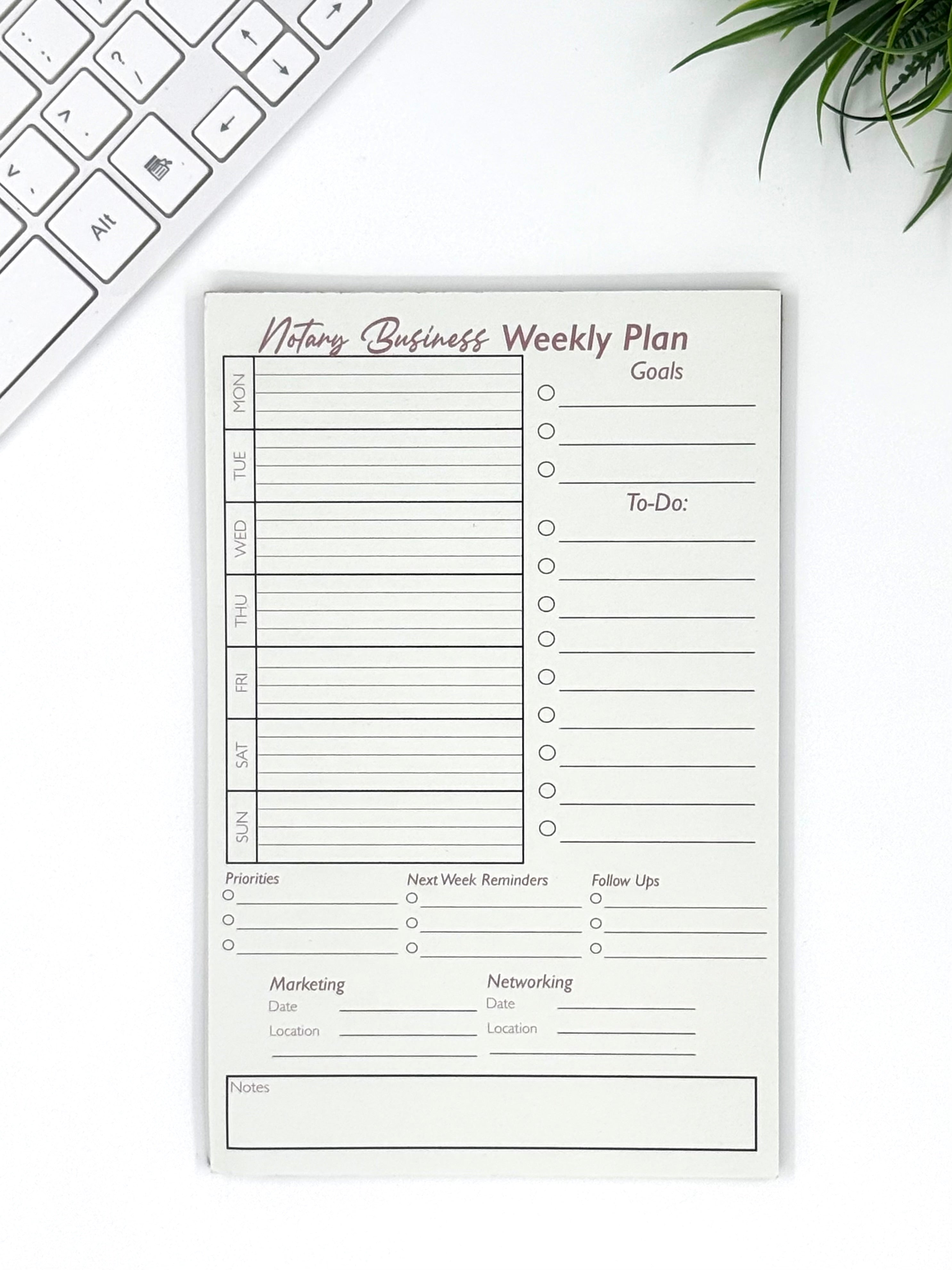 Weekly plan notepad for a business on a white surface with a keyboard and plant.