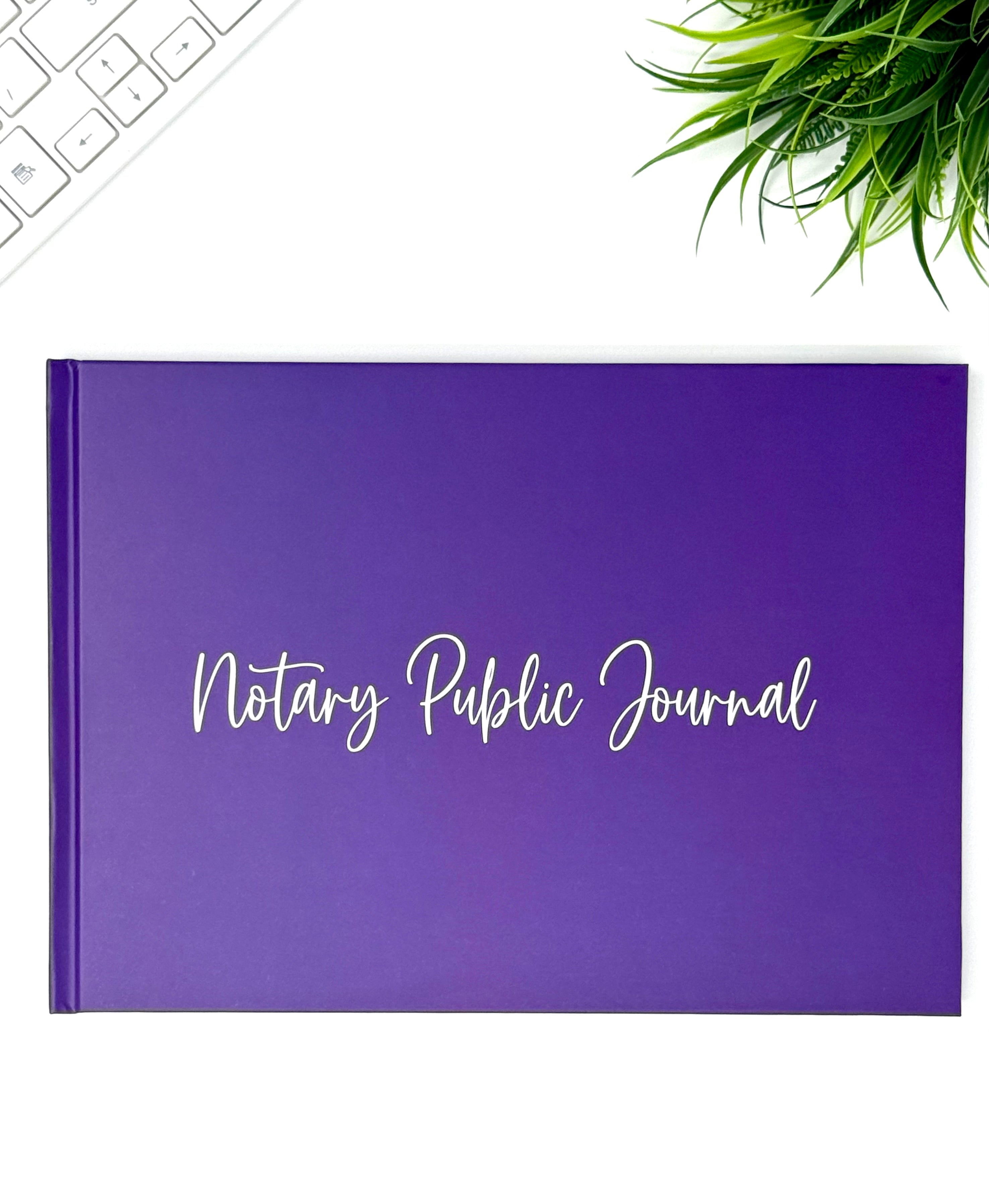 A purple Notary Public Journal with white cursive text on the cover, displayed on a white surface with a keyboard corner and green plant leaves visible.