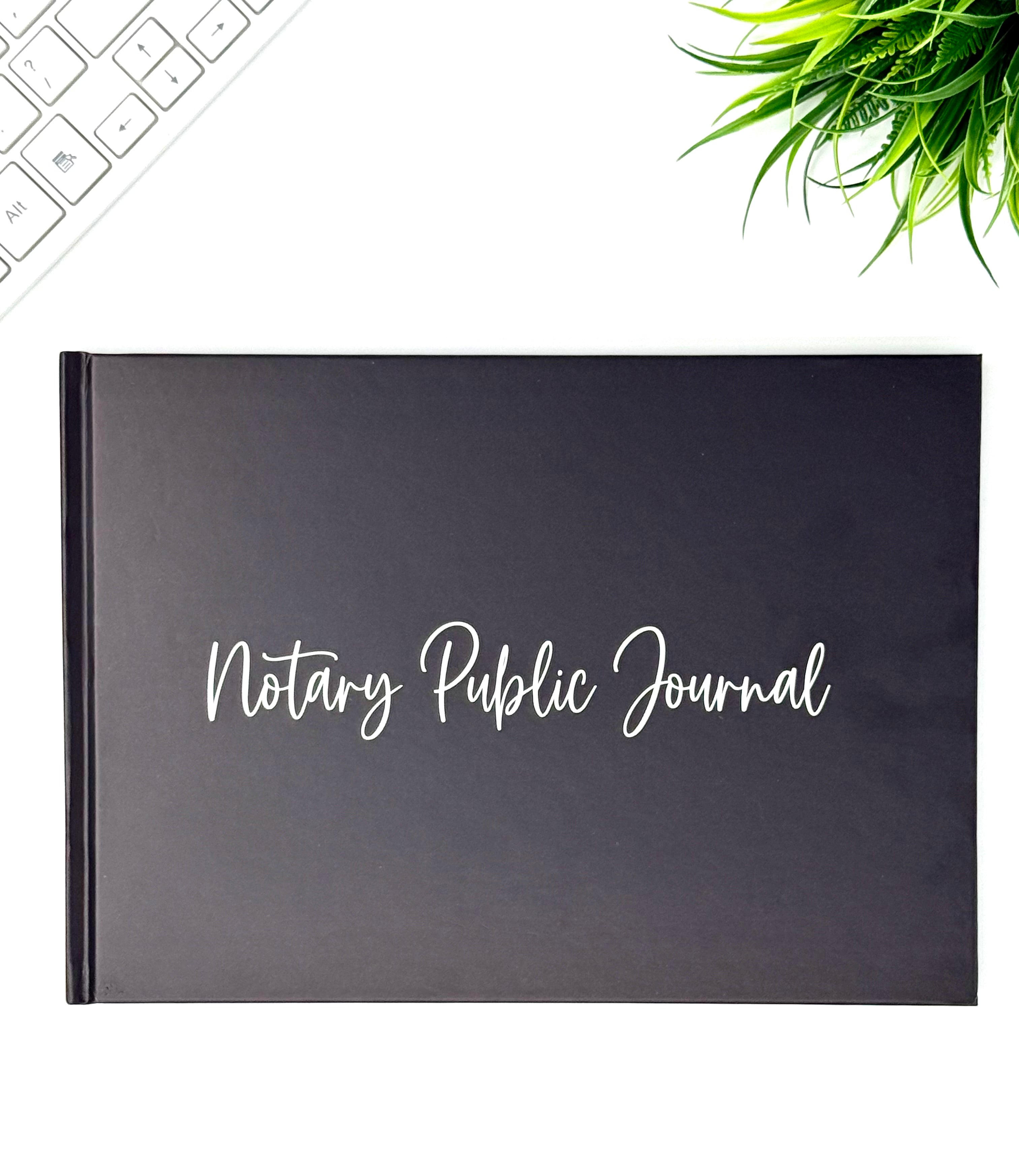 A black Notary Public Journal with white script lettering on the cover, displayed on a white surface with a keyboard corner and green plant leaves in the frame.