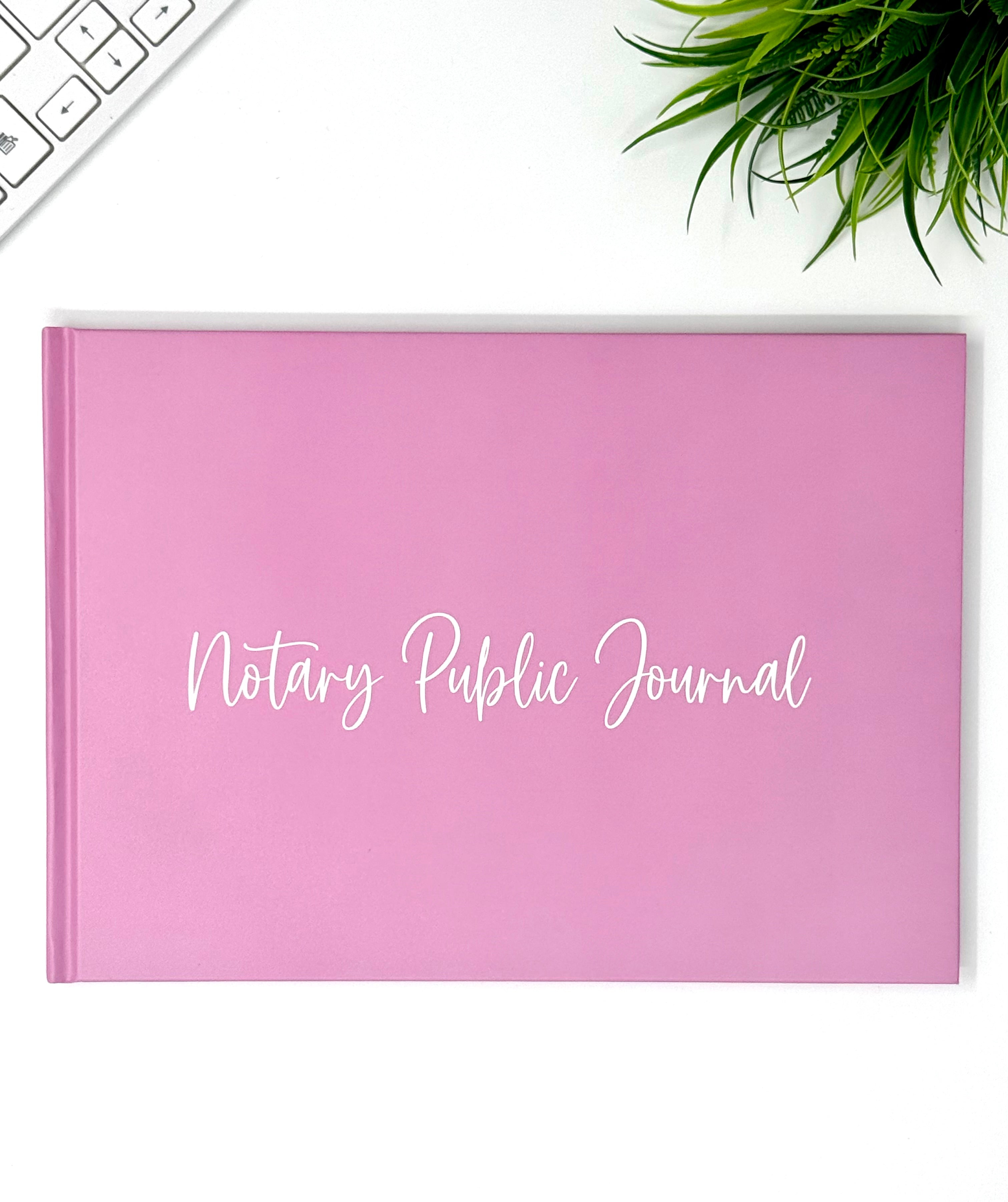 A pink hardcover Notary Public Journal with white script text on the front, displayed on a white desk with a keyboard corner and a green plant visible.