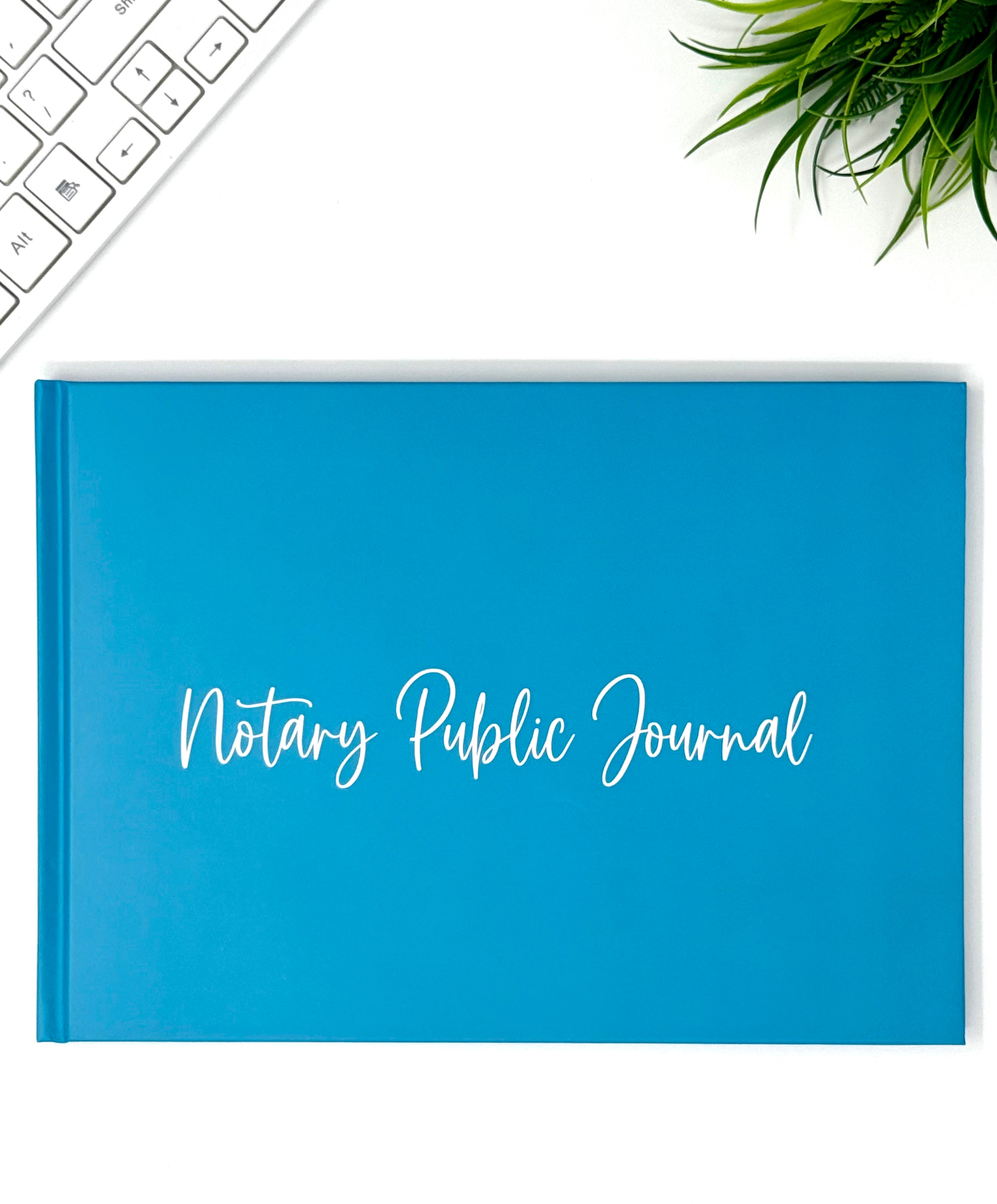 A bright blue hardcover Notary Public Journal with white script lettering on the front, photographed from above on a white desk with a keyboard corner and a green plant visible.