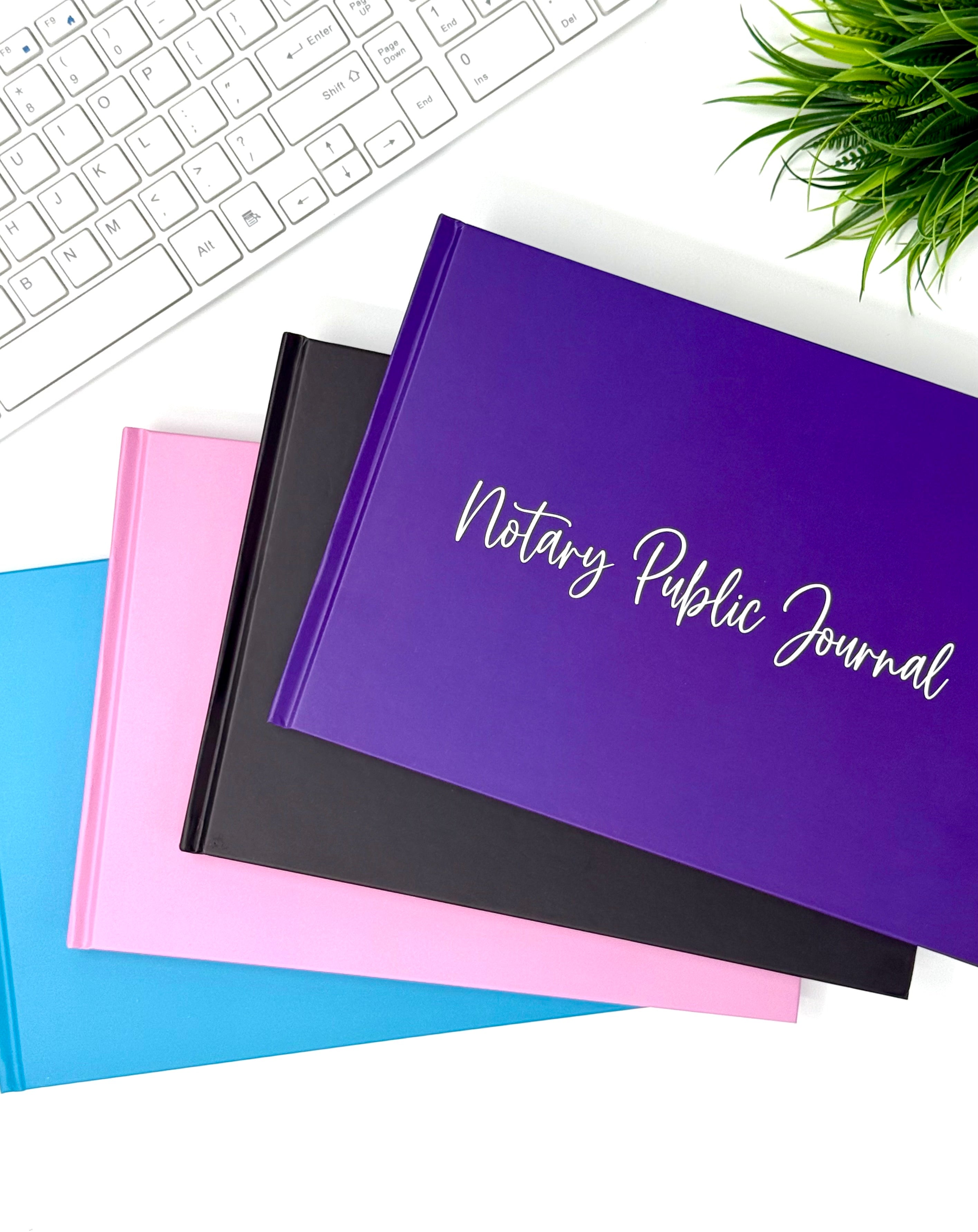 A stack of hardcover notary public journals in purple, black, pink, and blue, with the top purple journal featuring white script text reading 'Notary Public Journal.' The journals are arranged on a white desk with a keyboard, green plant, and small decorative object visible around them.