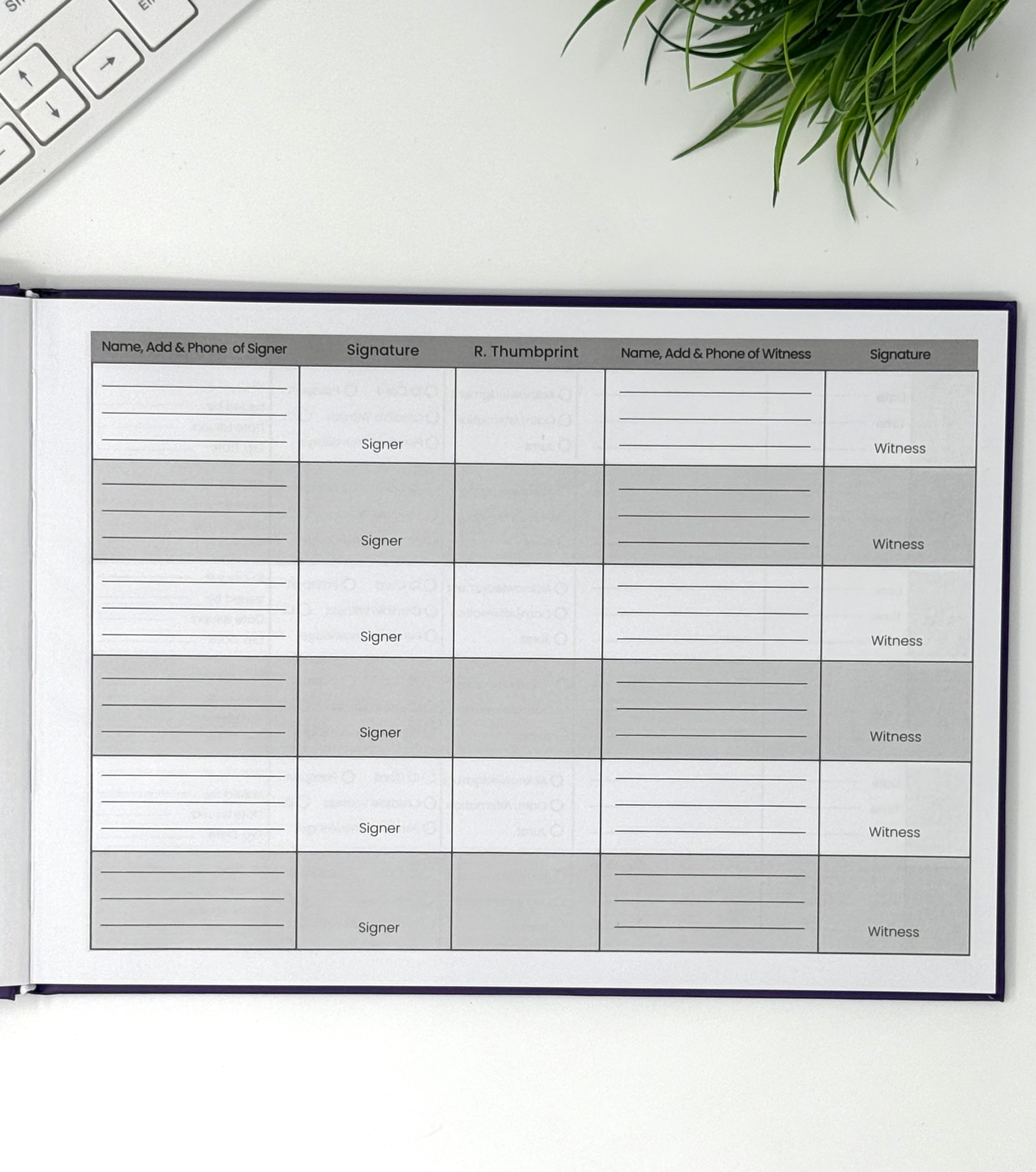 An open notebook or planner showing a printed witness statement form with columns for names, signatures, and thumbprints, photographed on a white desk with a keyboard corner and green plant leaves visible.