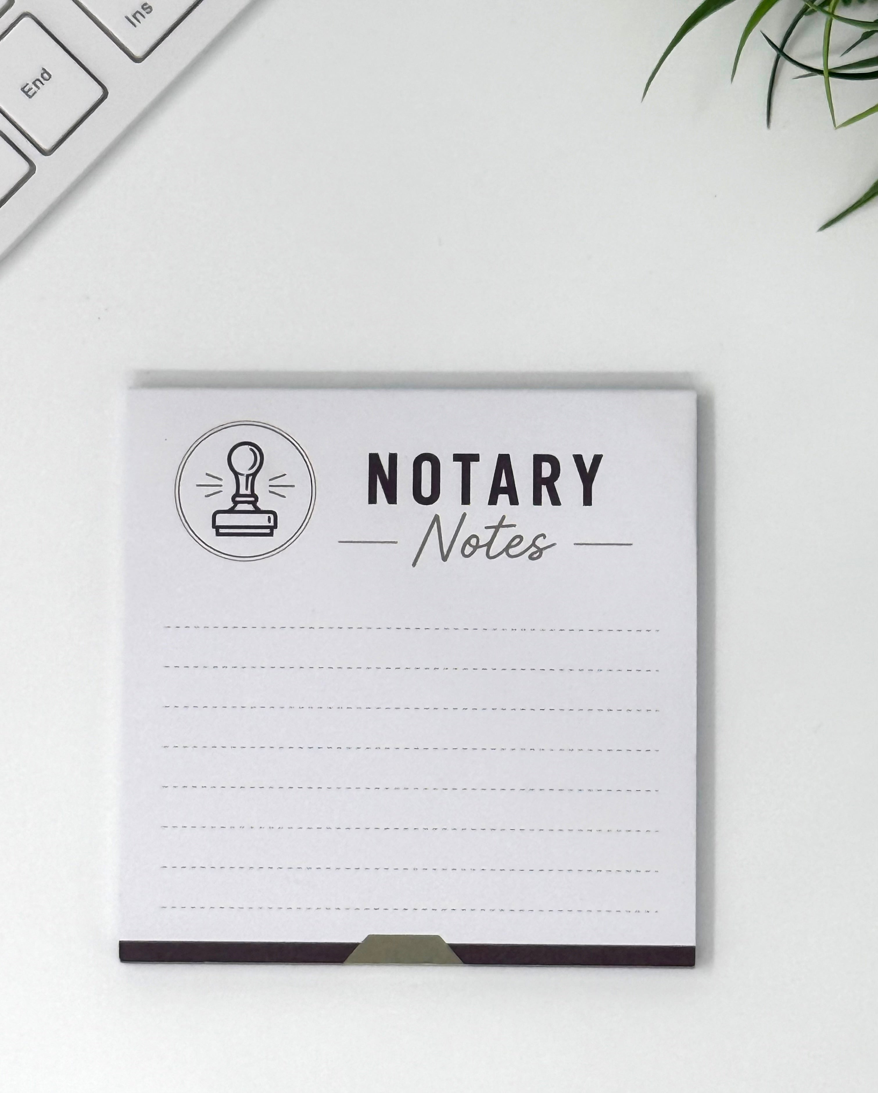 A square white notepad with the words 'NOTARY Notes' and a small stamp icon printed on the cover, photographed on a light desk surface with part of a keyboard and green leaves visible around it.