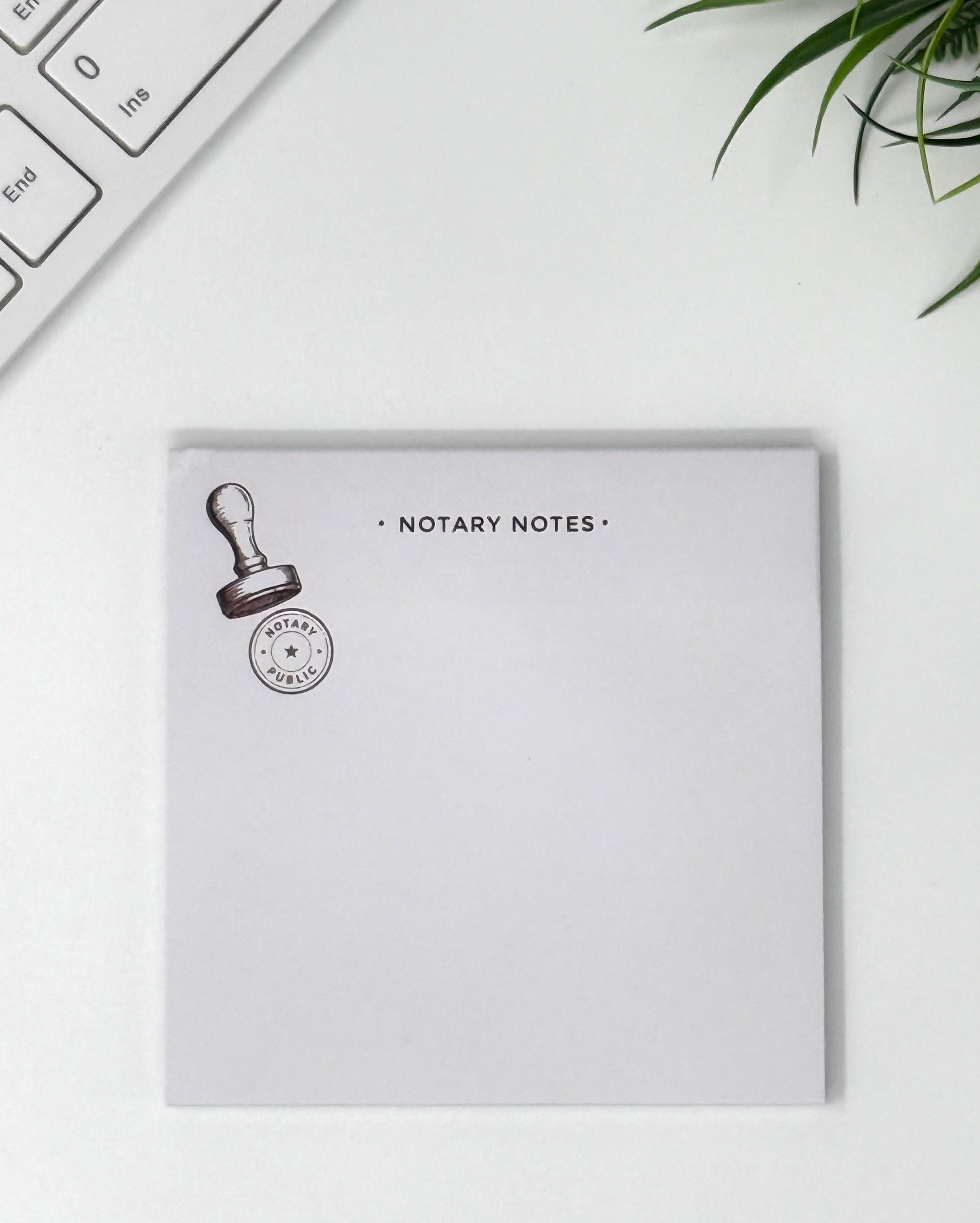A square white notepad labeled 'NOTARY NOTES' with a small stamp graphic, photographed on a light desk surface with part of a keyboard and green leaves visible at the edges.