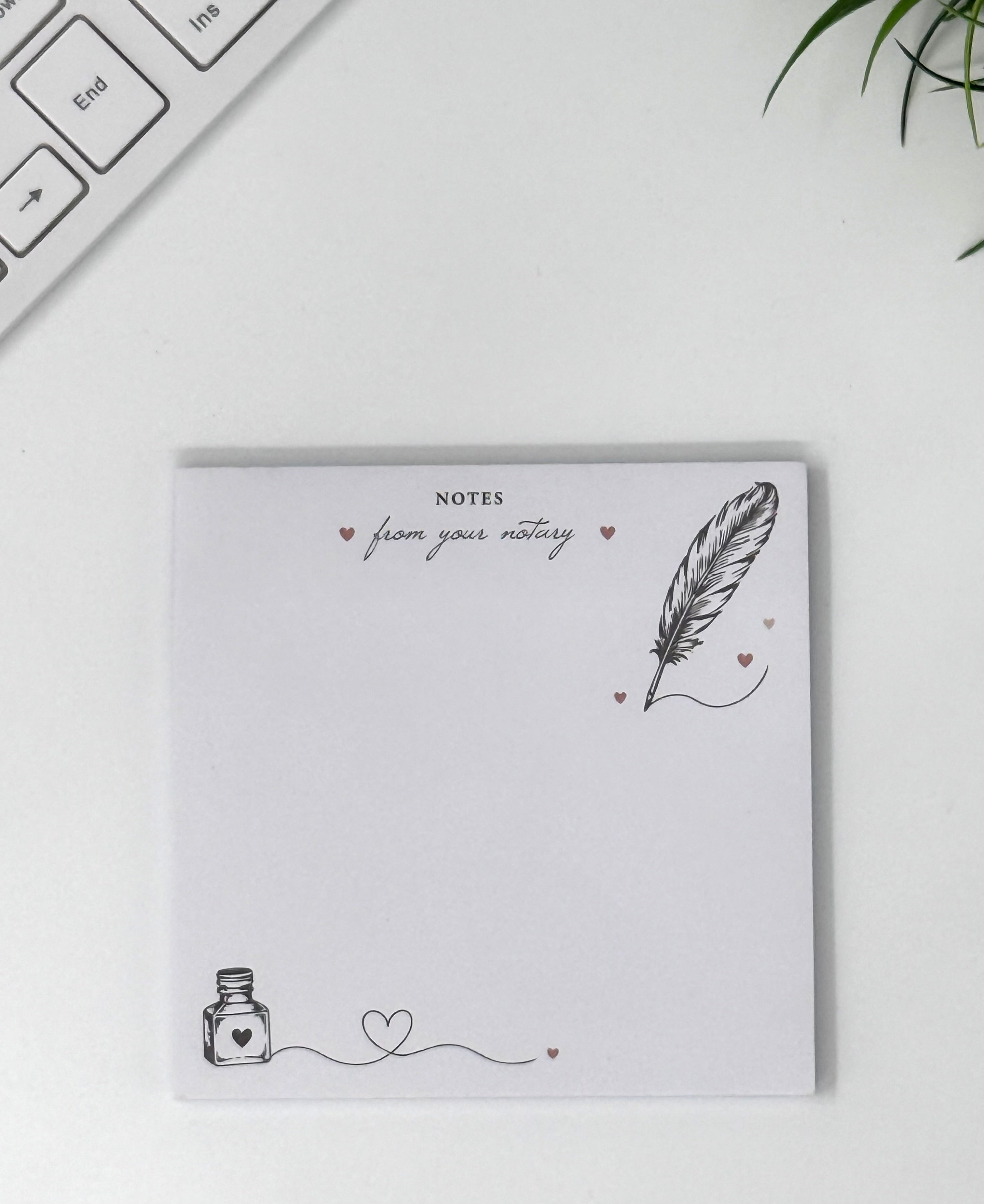 A square white notepad with a feather and ink bottle illustration, photographed on a light desk surface with part of a keyboard and a plant visible.