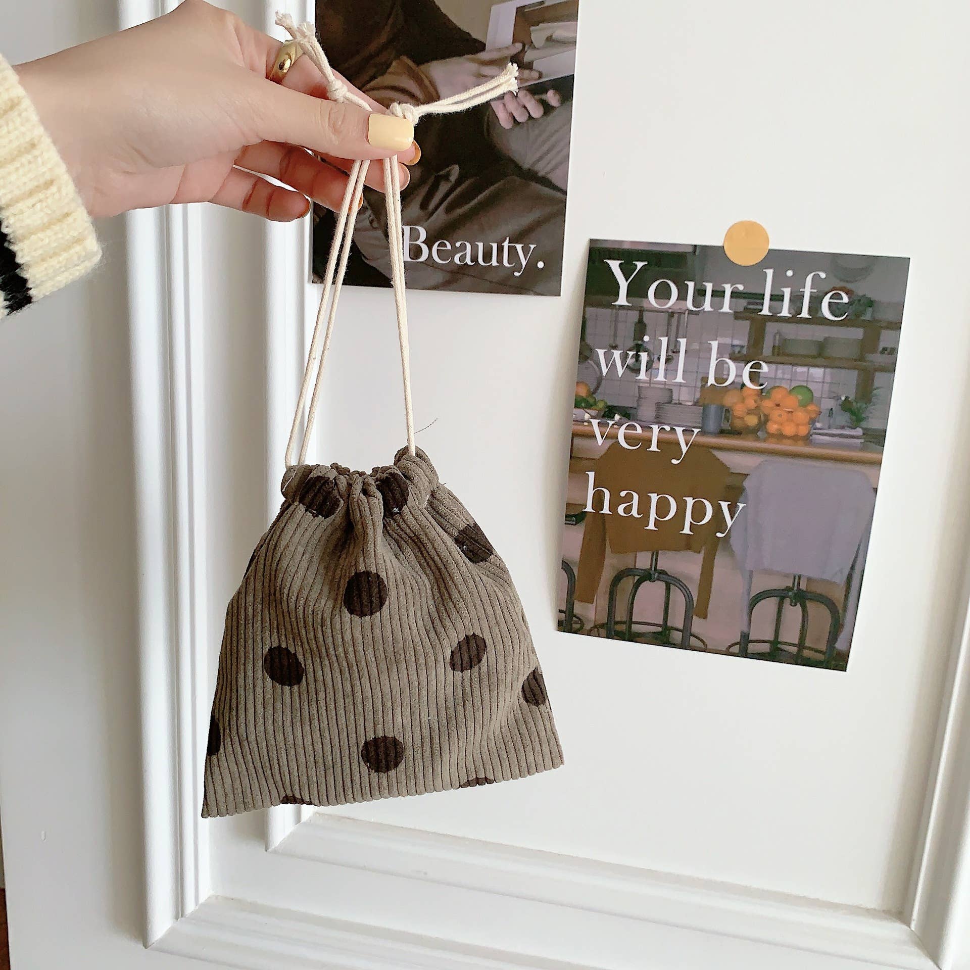 Small brown drawstring bag with black polka dots held by a hand against a white wall with framed pictures.