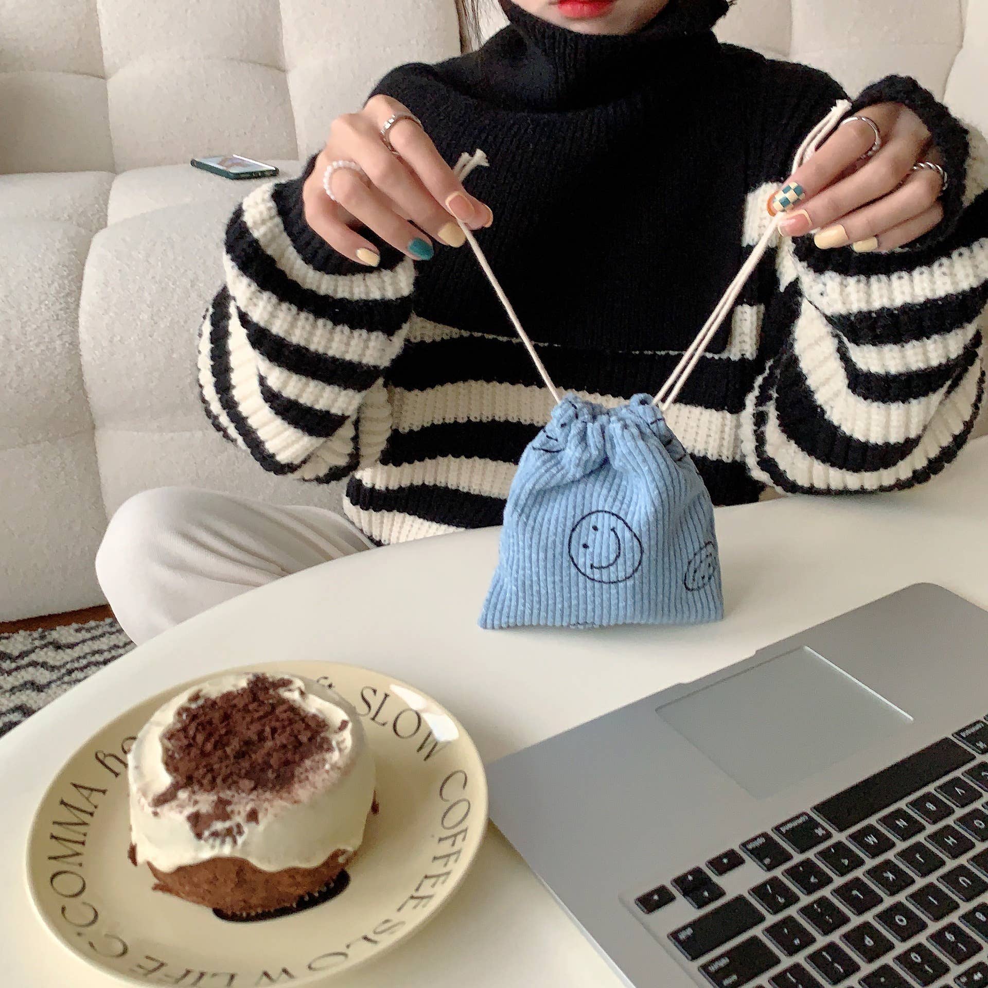 Person knitting with a blue drawstring bag on a table with a laptop and dessert.