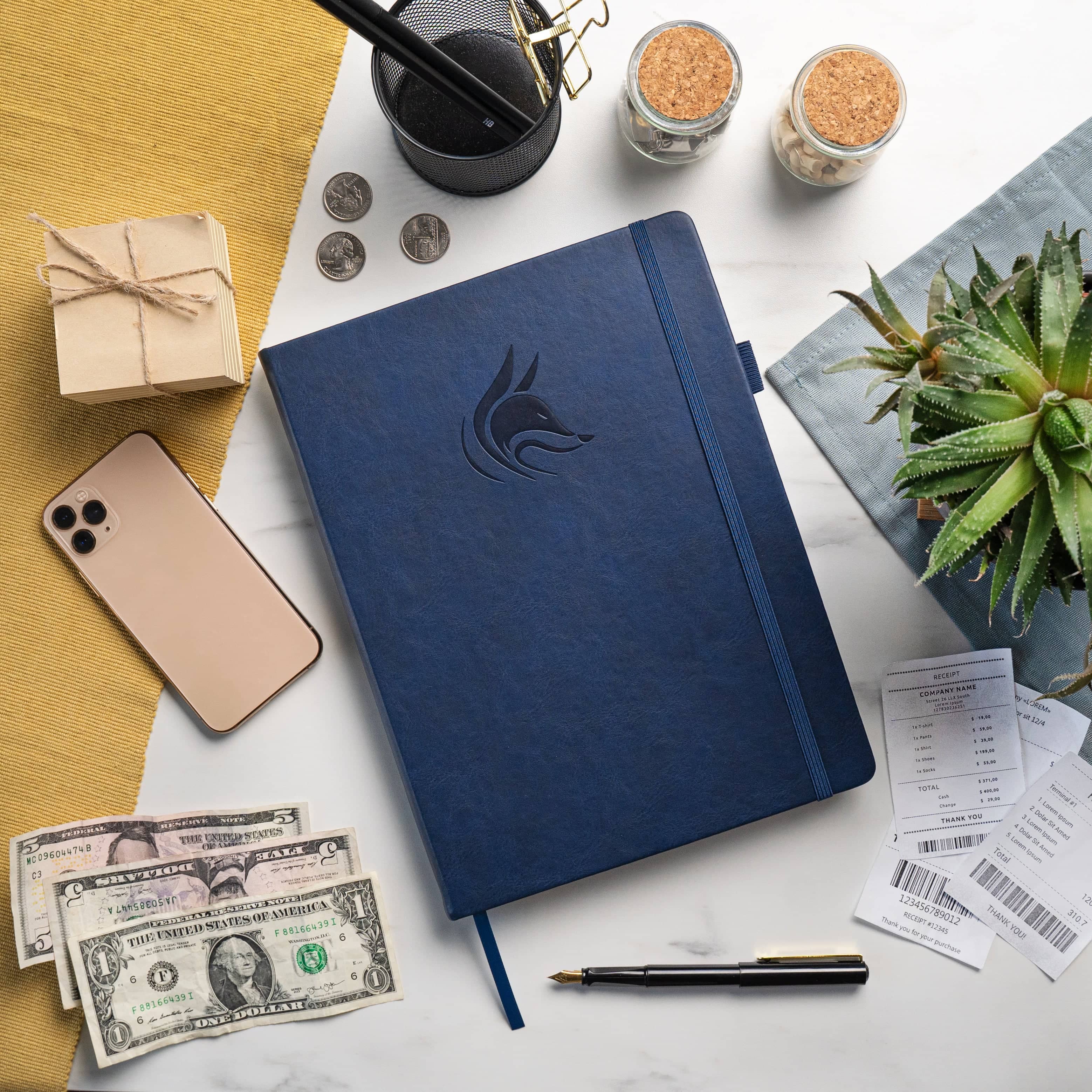A navy blue hardcover notebook with an embossed wolf logo, styled in a flat-lay arrangement on a white marble surface alongside a smartphone, cash, a pen, receipts, a small plant, and other desk accessories.
