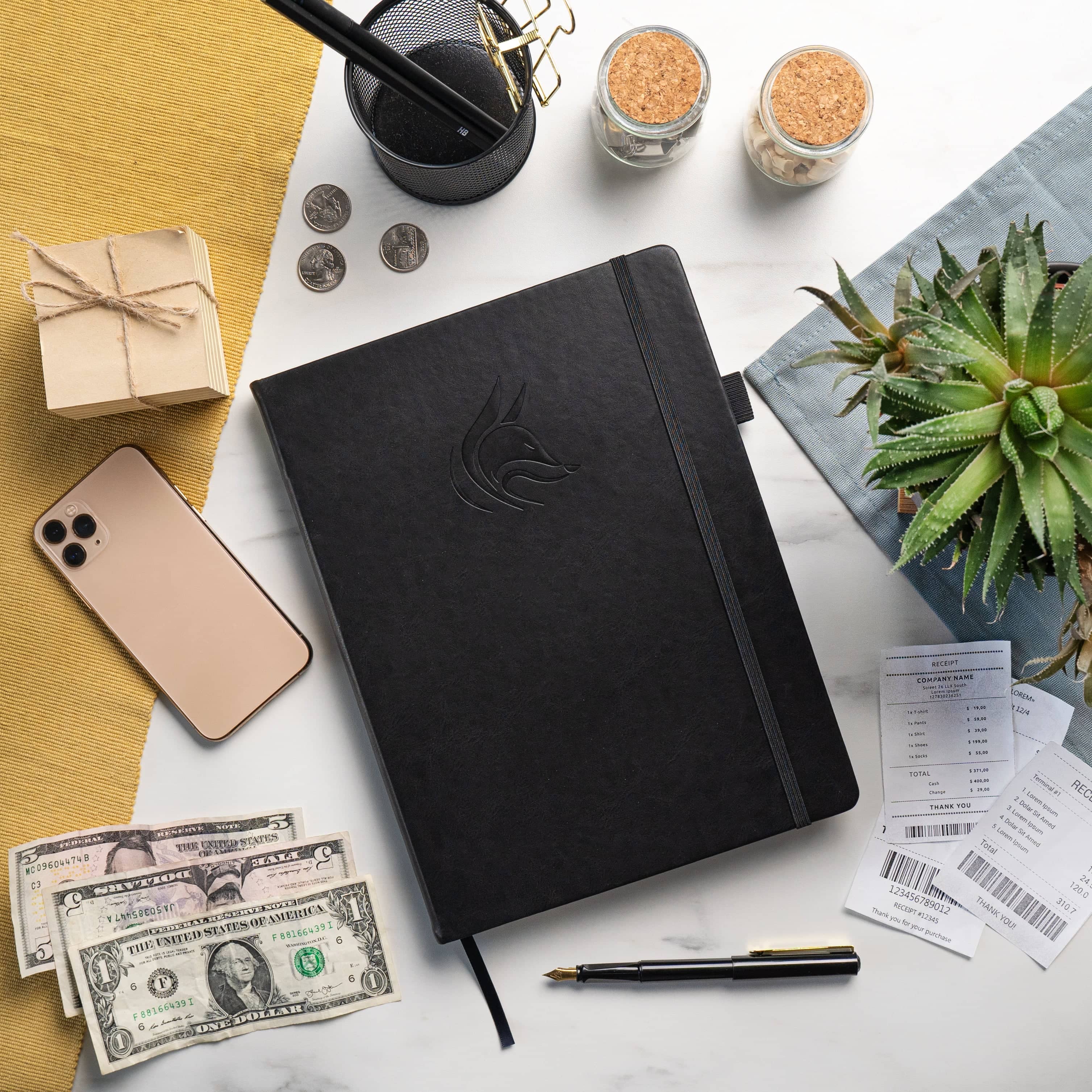 A black hardcover notebook with an embossed logo lies on a white marble surface surrounded by lifestyle props including a smartphone, cash, a pen, receipts, a small plant, and other stationery items.