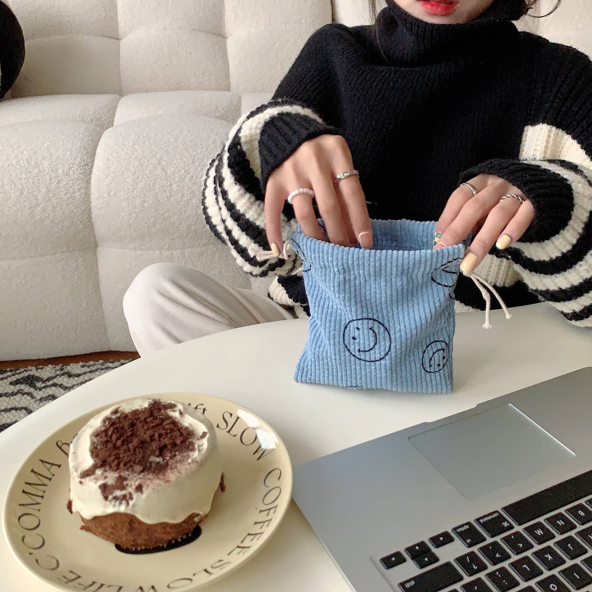 Person holding a blue drawstring bag with a smiley face design on a table with a laptop and dessert.