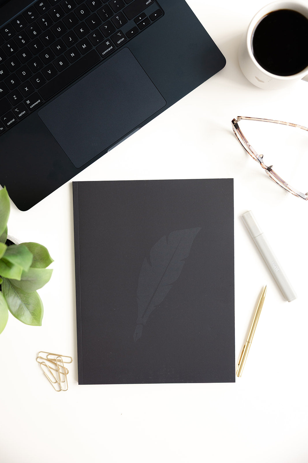 Black notebook on a white desk with a laptop, coffee cup, glasses, and pen.